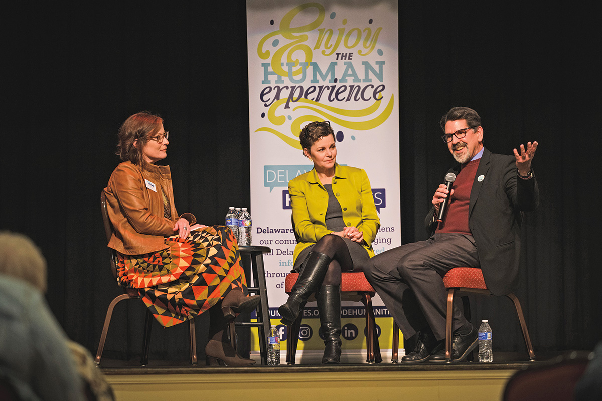 A group of three people sitting on stage. The person on the left and in the center are listening to the person on the right, who is speaking and addressing the audience. A poster behind the people says human experience. 