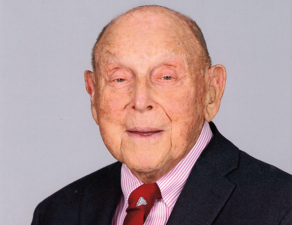 An older person with fair skin and very little hair, wearing a business shirt, tie, and jacket, smiles at the viewer.