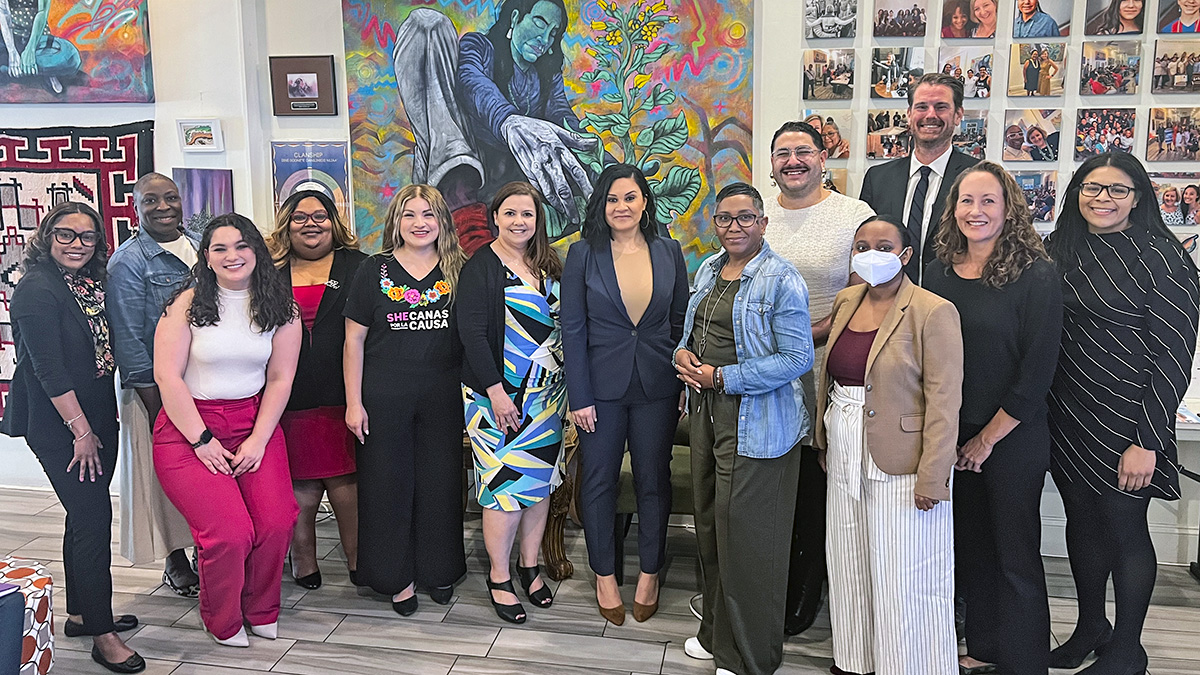 A group of young people of various races pose together in front of a large, colorful painting. They face the viewer and smile.