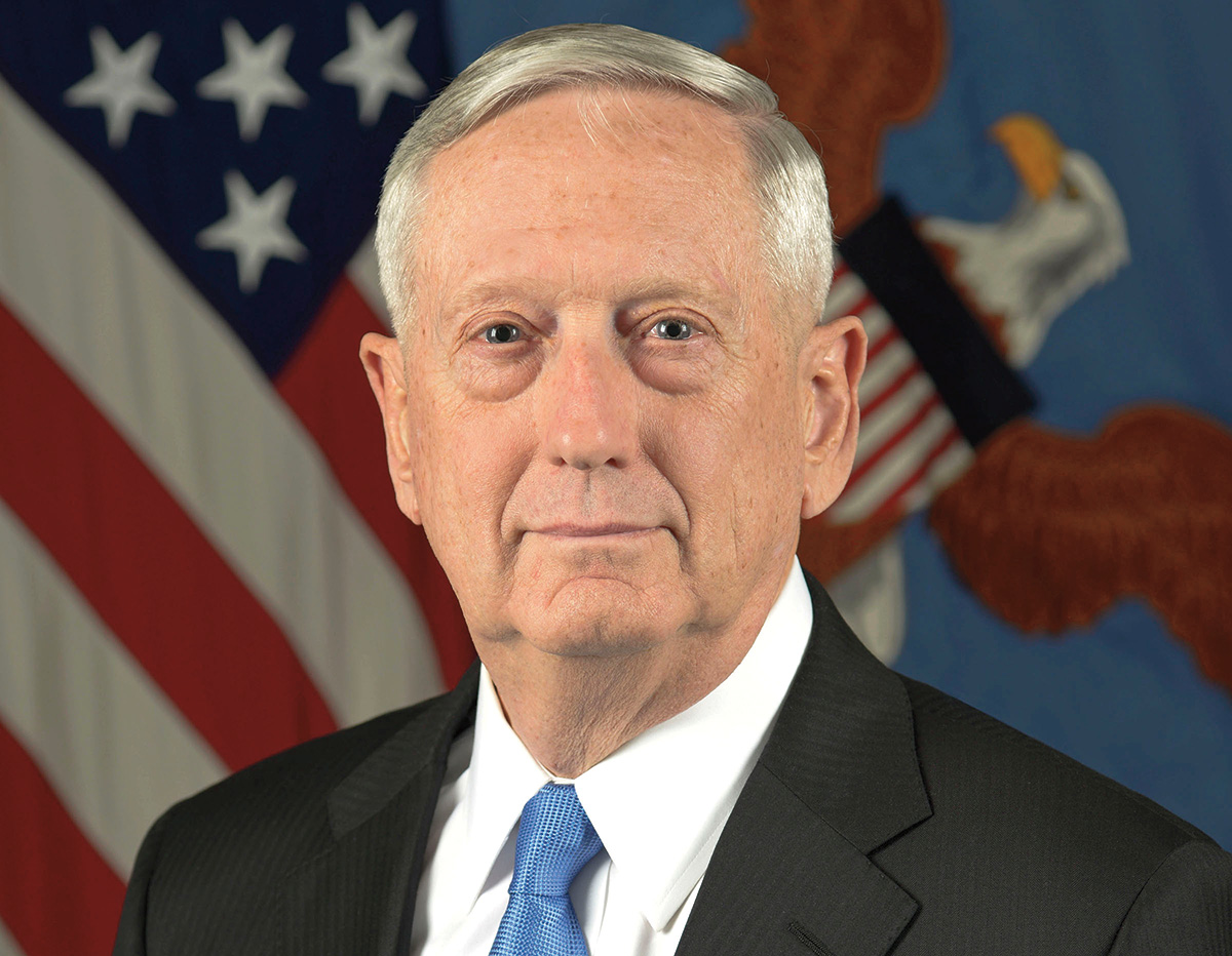 An individual with light skin and gray hair, wearing a business suit, smiles at the viewer. The American flag and the flag of the Secretary of Defense are in the background.