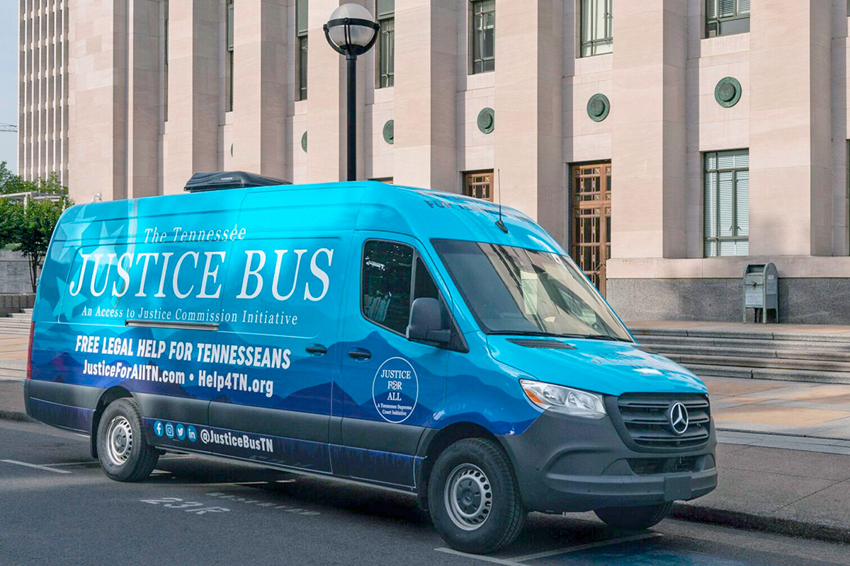 A blue cargo van is parked outside of a municipal building. The van's sides are covered in text that reads, "The Tennessee Justice Bus: An Access to Justice Commission Initiative. Free Legal Services for Tennesseans, JusticeForAllTN.com, Help4TN.org.