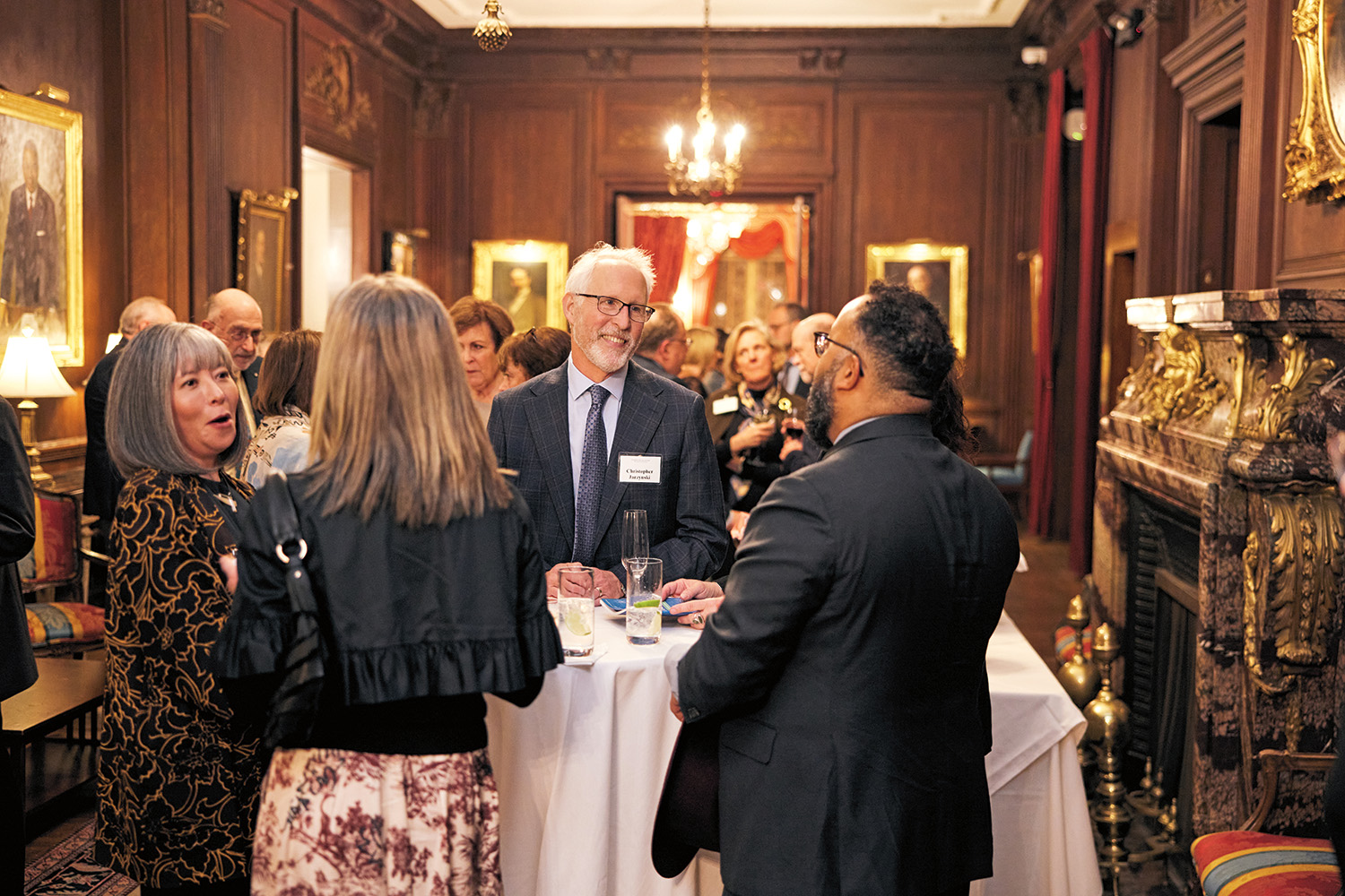 Formally dressed people are in conversation in a wood-paneled room with chandeliers and gold-framed portraits. 