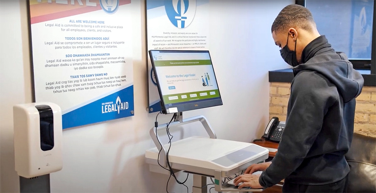 A person stands at kiosk station typing into a computer. The person has short dark hair and wears a black facemask and turtleneck. A welcome banner in multiple languages in large print with the Mid-Minnesota Legal Aid logo is posted on the wall and a phone sits on a desk with a chair.