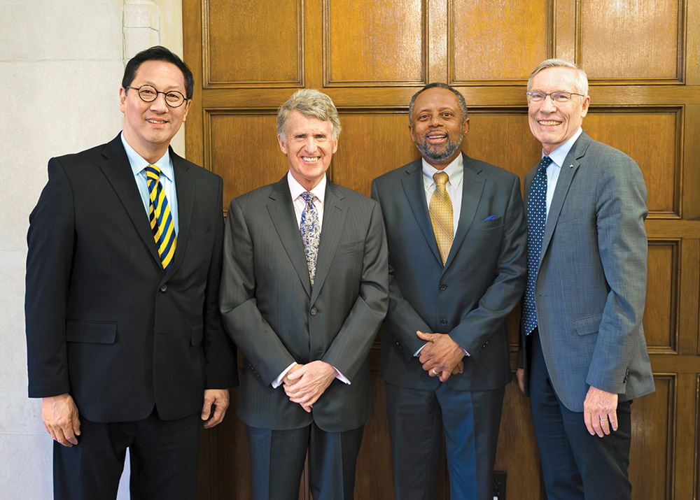 Four formally dressed men pose with a wooden wall behind them. 