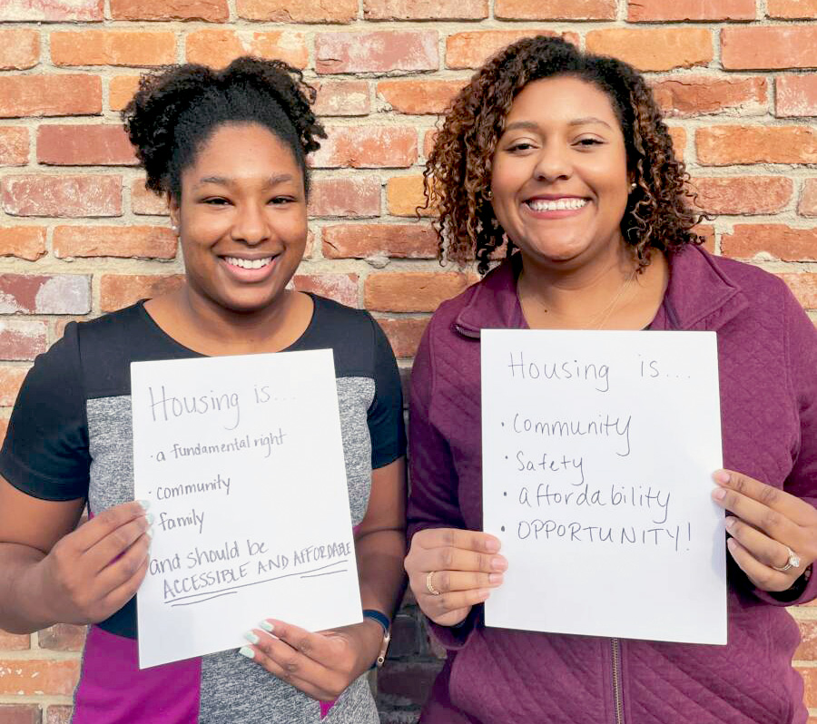 Two people stand together, holding signs that read "Housing is a fundamental right," “Housing is community,” and “Housing should be accessible and affordable.” Both have brown skin and long dark hair. They face the viewer and smile.