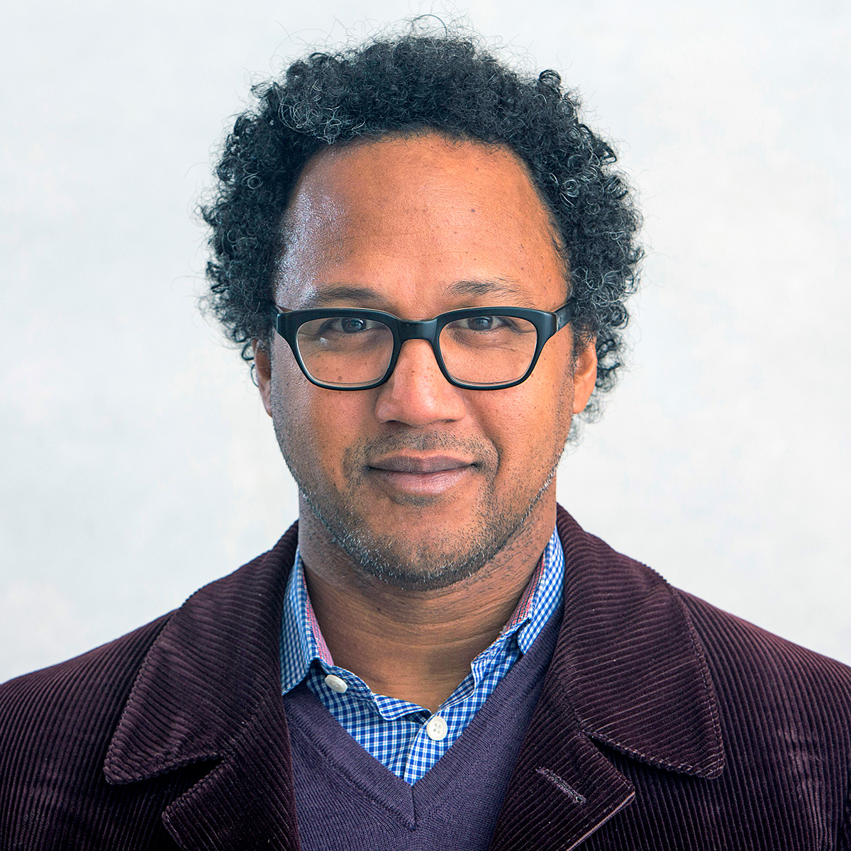 A headshot of André Fenton. Fenton has brown skin and black curly hair. He wears black glasses, a burgundy blazer, a blue dress shirt, and a gray sweater. He faces the camera and smiles.