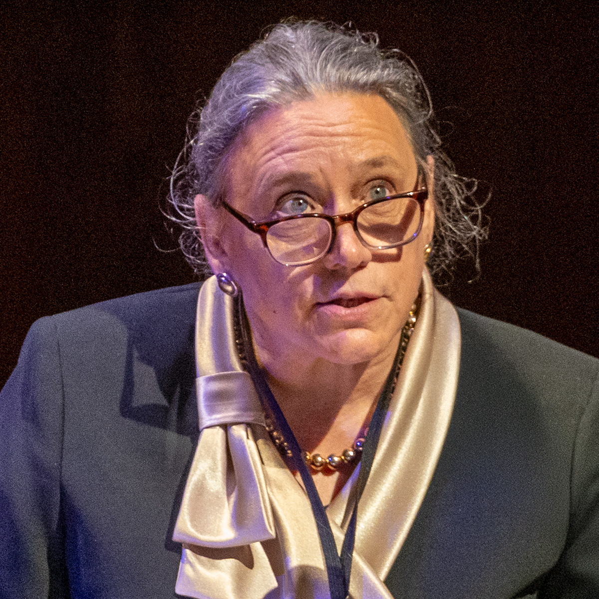 A portrait of Laurie L. Patton. Patton has pale skin, straight long gray hair, and blue eyes. She wears business attire and stands in front of a black background. 