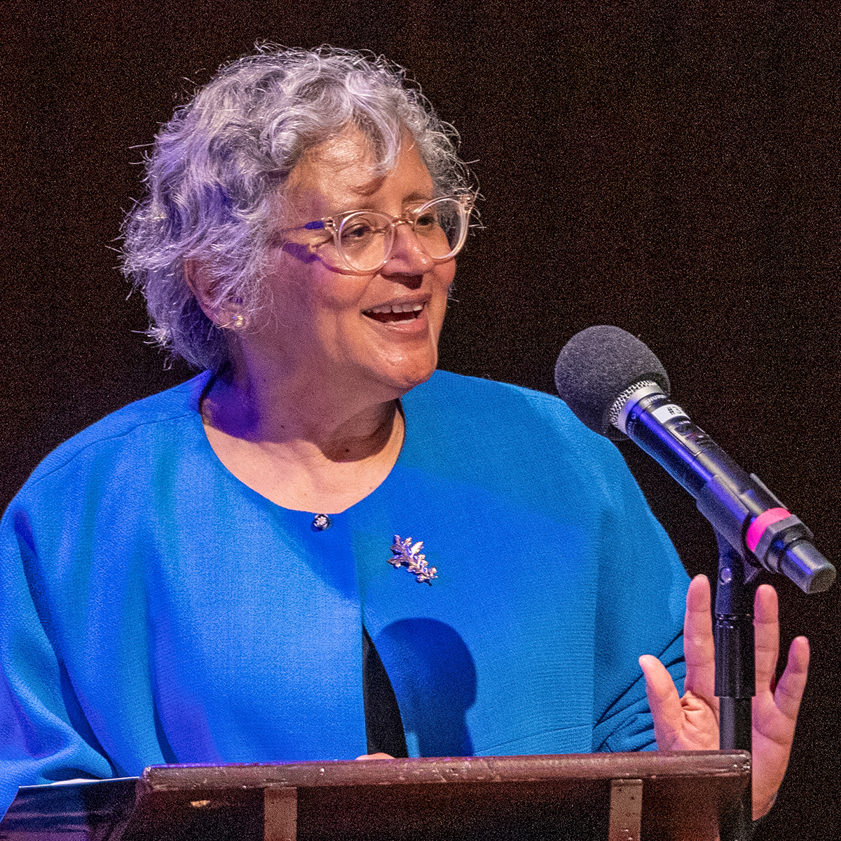 A photograph of Cecilia Conrad, a woman with wavy gray hair, wearing glasses and a blue top. She is speaking into a microphone and addressing an audience not shown. 