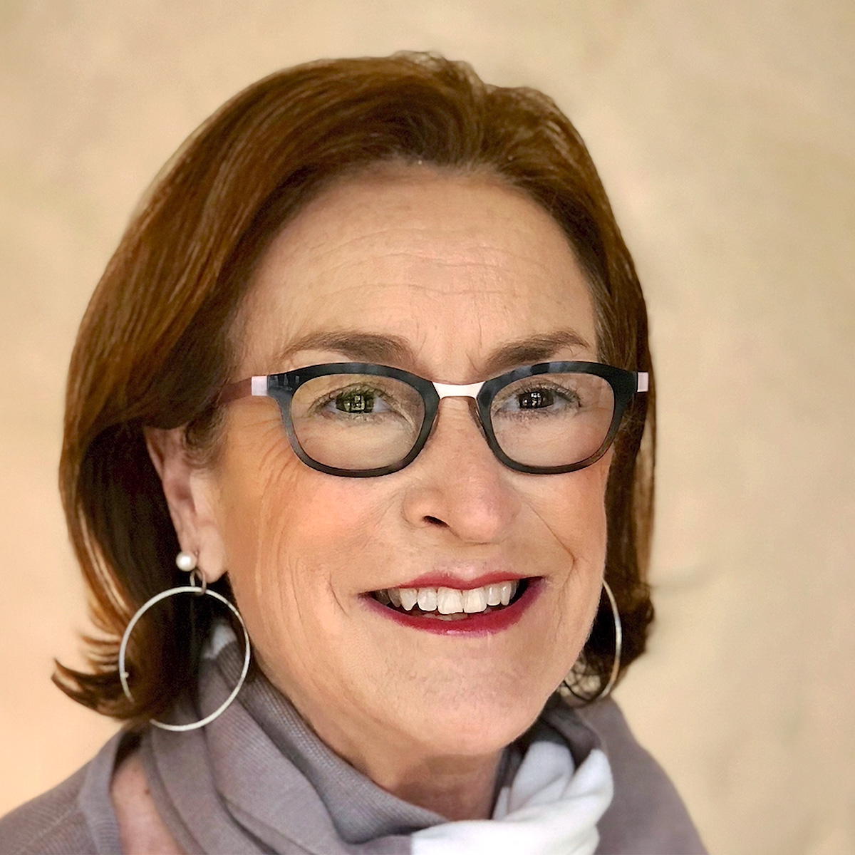 A photo of Cristine Russell, a light-skinned woman with reddish brown hair, wearing black frame glasses, silver hoop earrings, and a gray and white scarf.  She is smiling at the viewer.