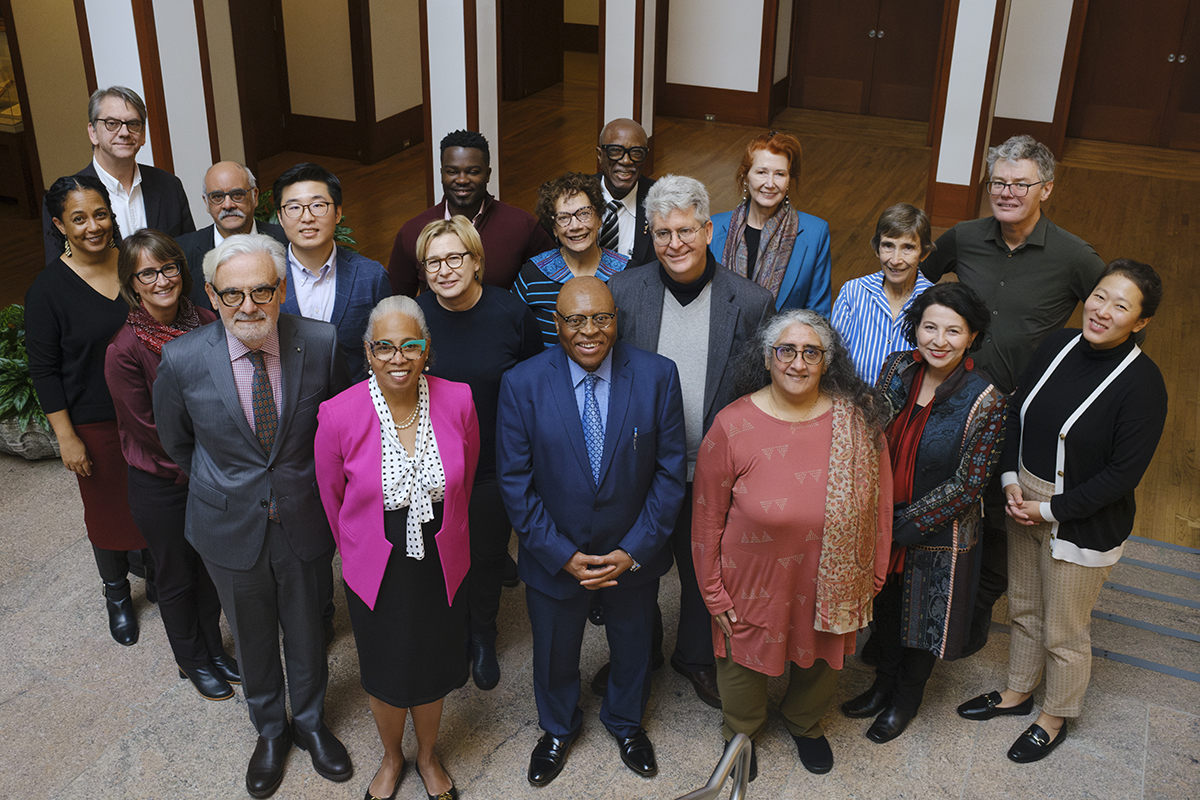 A group of nineteen scholars stand in the lobby of the American Academy of Arts and Sciences for “The Global Quest for Educational Equity” conference. Their appearances vary. They all wear professional clothing. All smile and face the viewer.