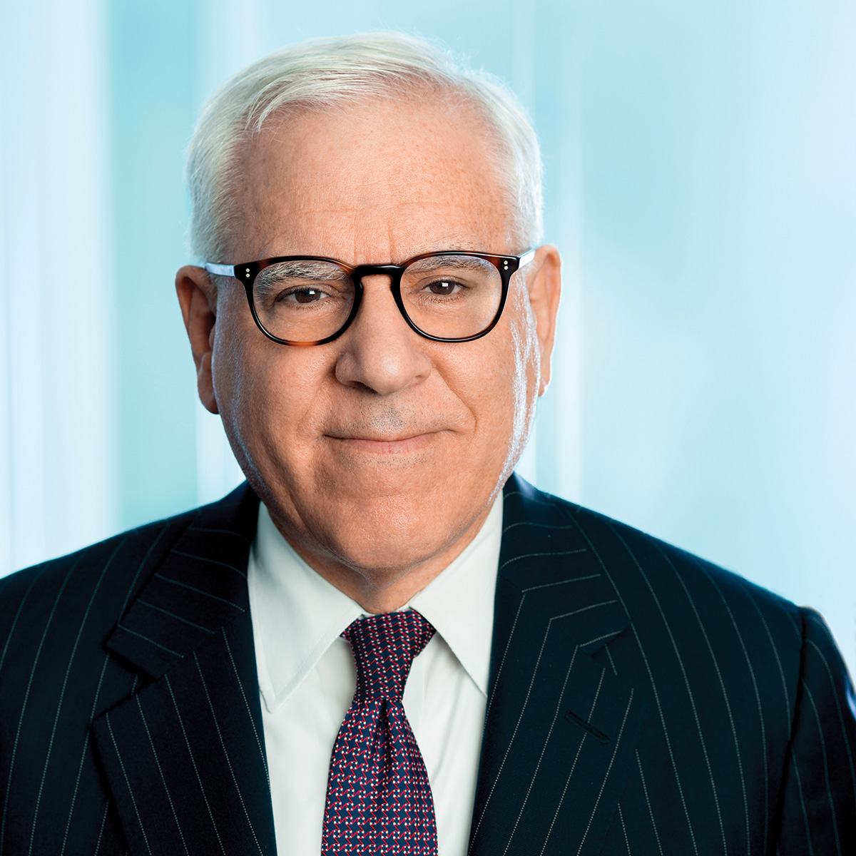 A headshot of David M. Rubenstein. He faces the camera and smiles. He has pale skin and short white hair. He wears plastic glasses, a blue and red tie, a white collared shirt, and a blue pinstriped suit. Photo courtesy of David M. Rubenstein.