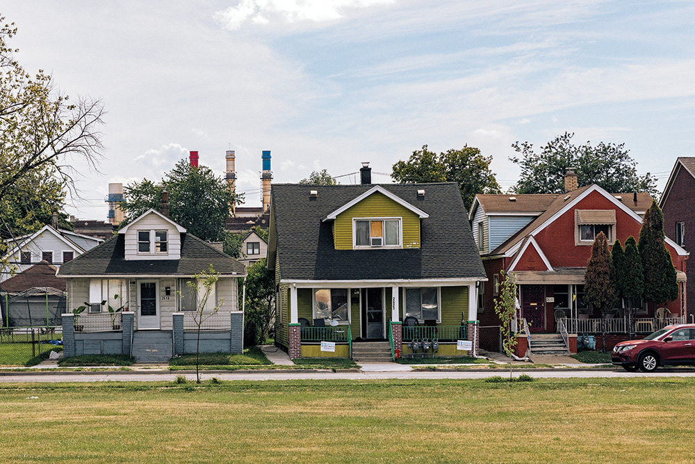 Homes across from a well-manicured grass field, with smokestacks in the background. 