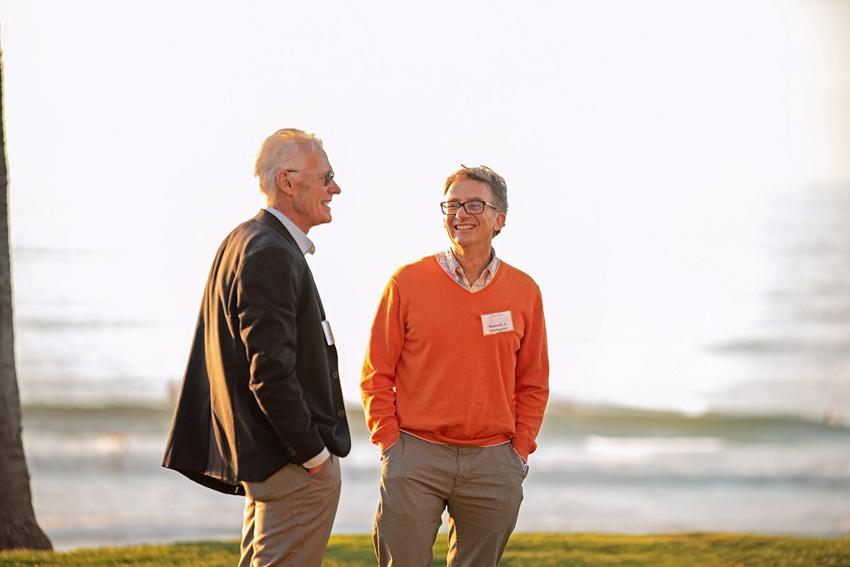 Two adults, standing outside with the ocean in the background, smile at each other.