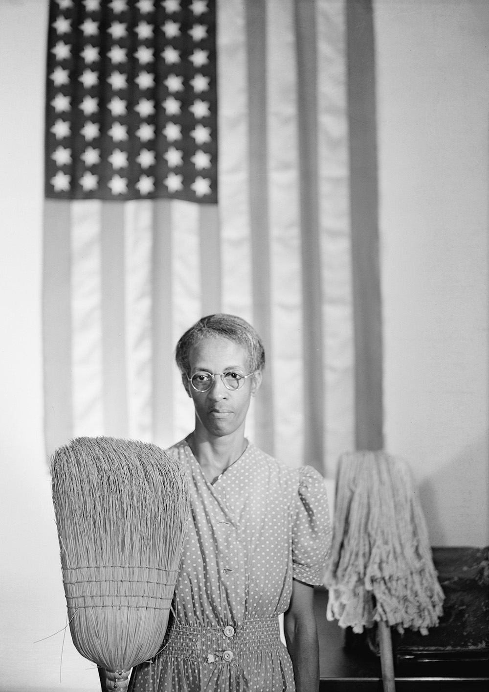 A person with dark skin standing in front of the American flag, holding a broom and a mop.