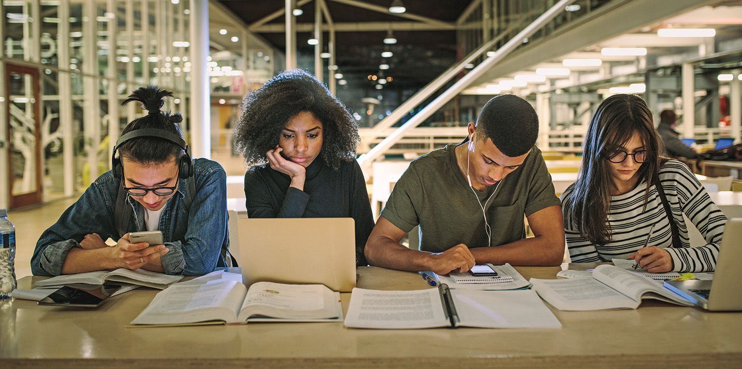 Four college-age students study together at a table covered by their laptops, phones, and books. Two students are engaged on their phones, another student looks at their laptop, and the last student is writing in a notebook.