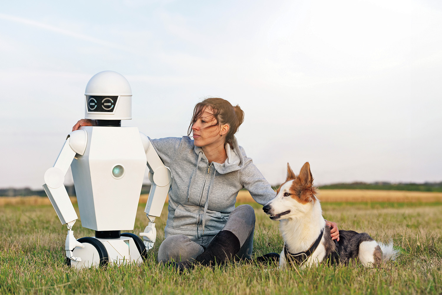 A person sitting in a field with their arms around a dog on their right and a robot on their left.