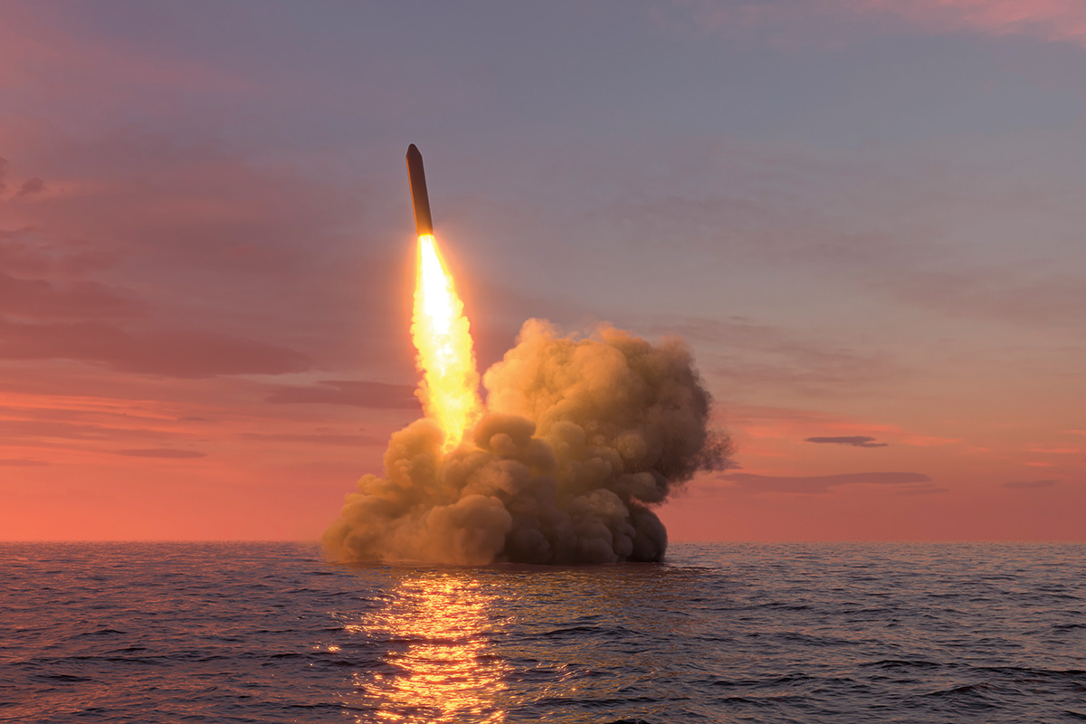 A ballistic missile launches from the ocean, with flames shooting upward and smoke trailing down. The ocean is calm with small ripples. The missile is set against a pink and blue sky.