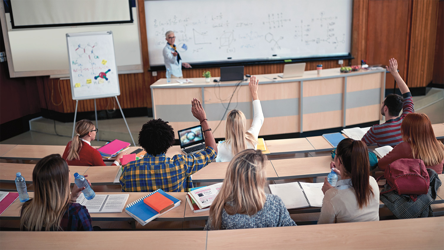 A group of students seated in a lecture hall face the instructor, who is standing in front of a white board with scientific notations and diagrams. Three of the students have their hands raised. 