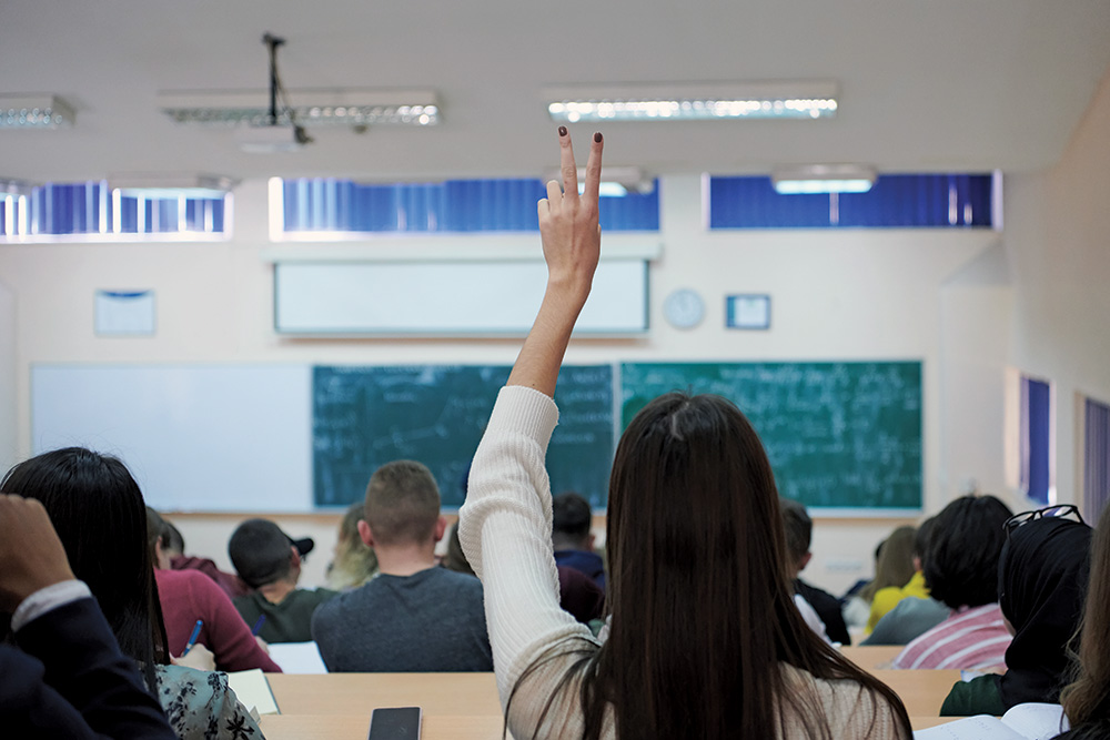 A view from the back of high school students facing a blackboard in a classroom. A student in the center of the class raises their hand.