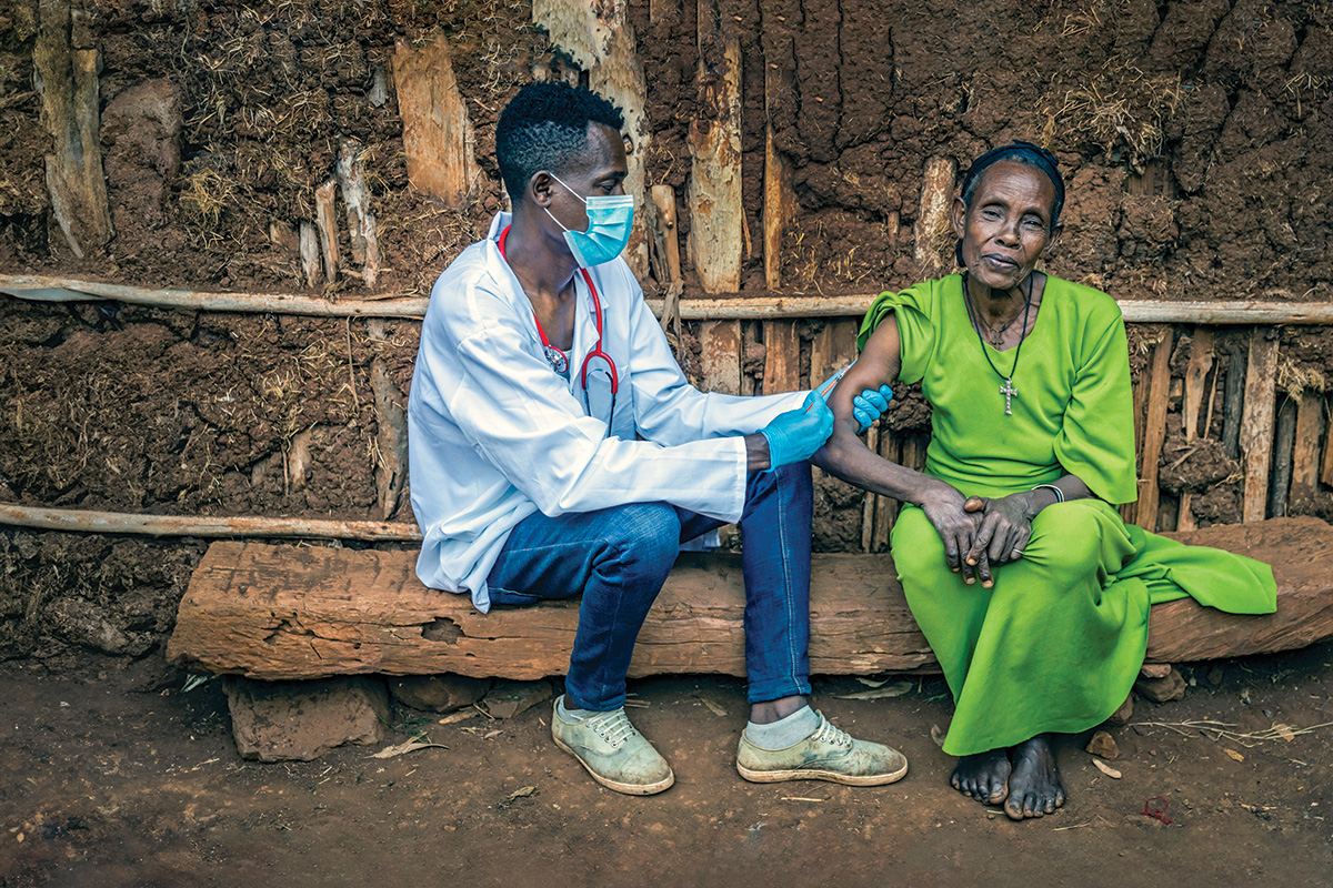 A middle-aged man with short black hair and brown skin, wearing surgical gloves, a mask, and a stethoscope, inoculates an older woman with gray hair, wearing a black headband and a bright green dress, and sitting on a wooden bench in front of a wall made from earth and wood. The man faces the woman, and the woman faces the viewer.  