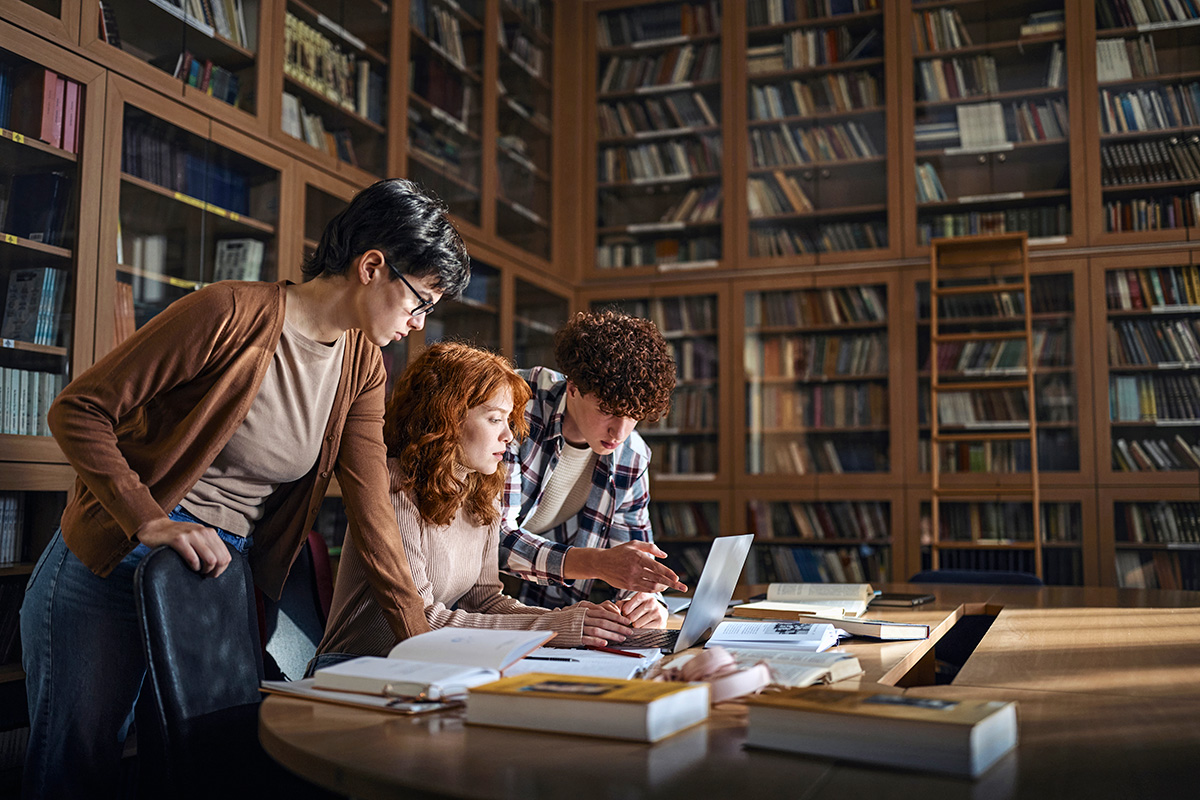 Three young individuals are looking at a laptop in a library setting, surrounded by books and study materials.