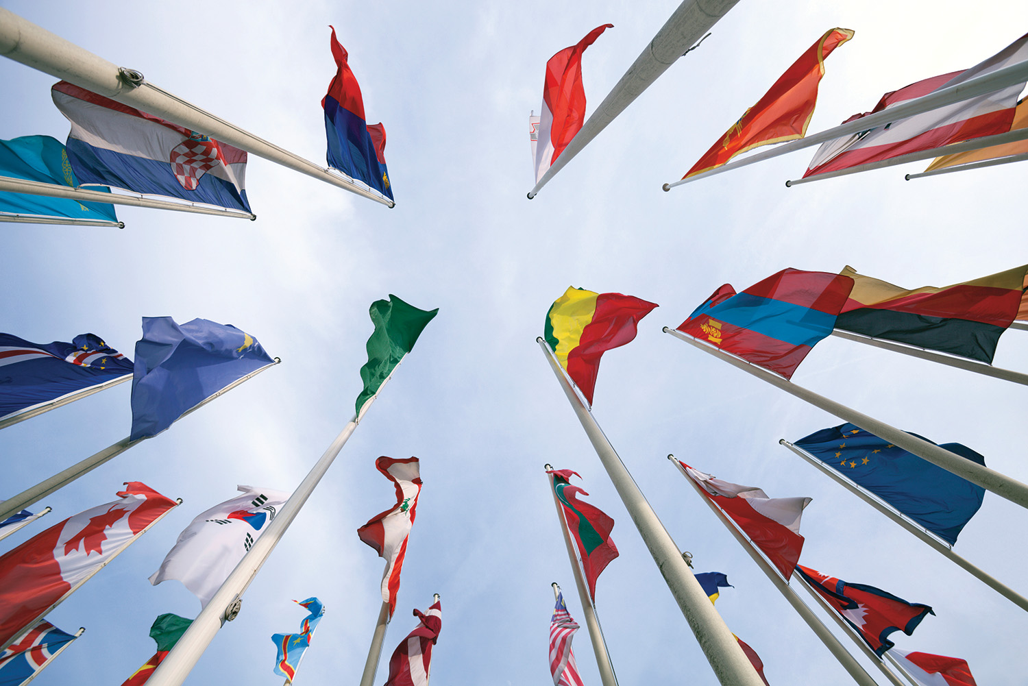 More than a dozen flags from various countries are attached to tall flagpoles. They are viewed from the ground up against a blue sky. 