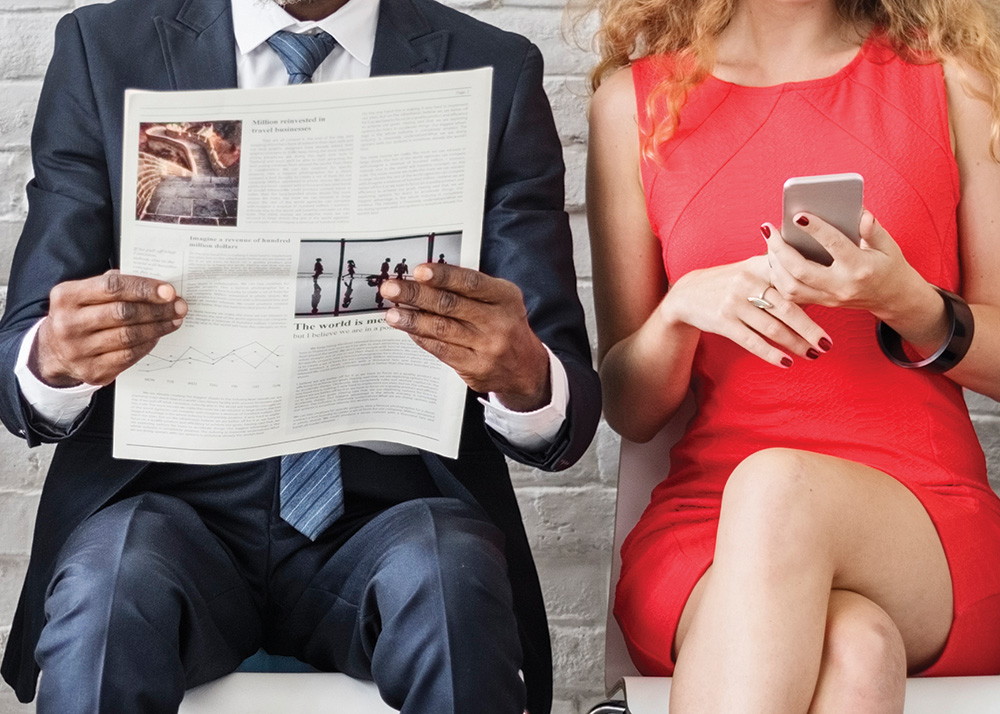 Three individuals sitting on a couch. The person on the left is using a laptop. The person in the middle is reading a newspaper. The person on the right is reading their phone.