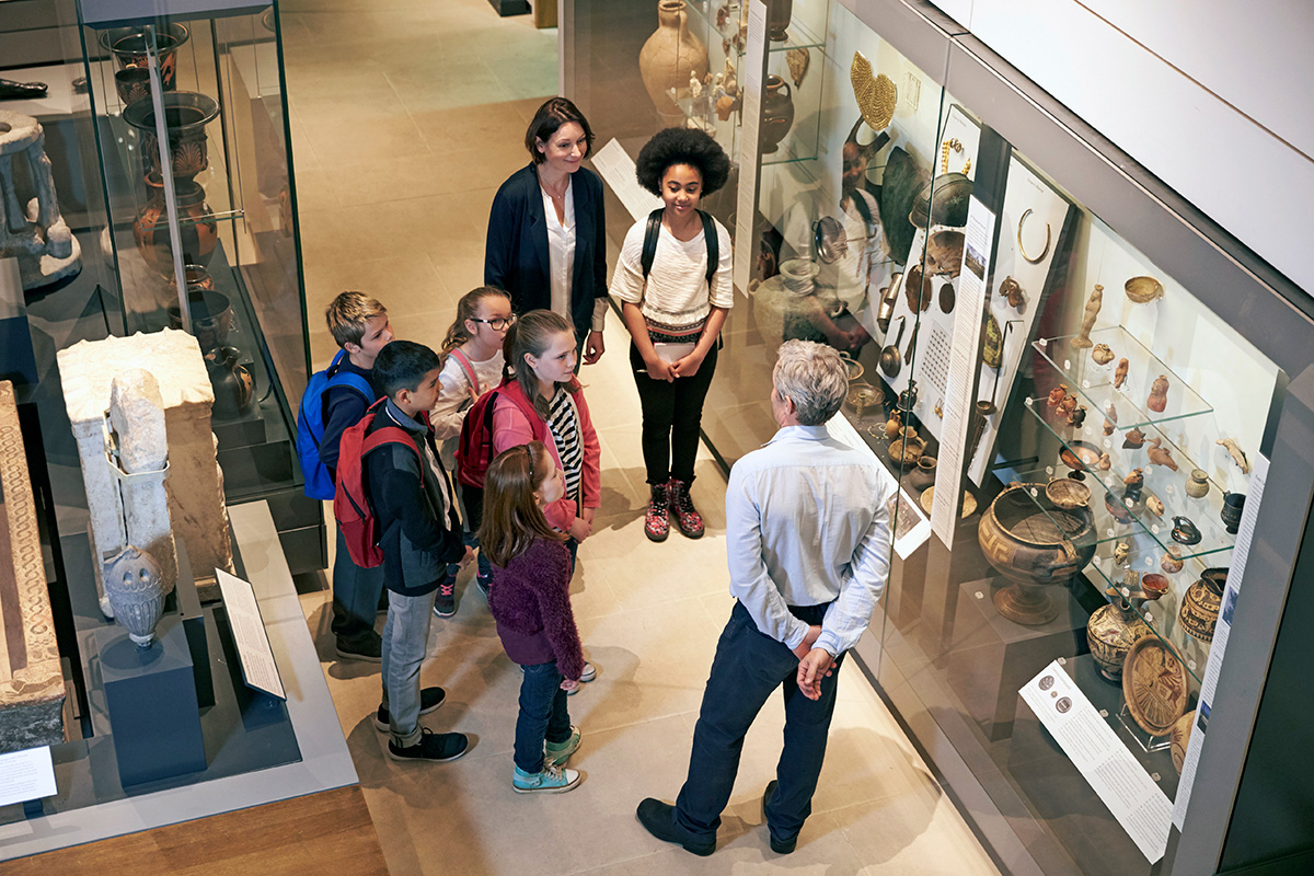 A group of children, accompanied by two adults, are standing in a museum gallery, listening to an older man, who is explaining the exhibits of artifacts in the display cases around them. The cases feature ancient pottery, sculptures, and other historical artifacts.