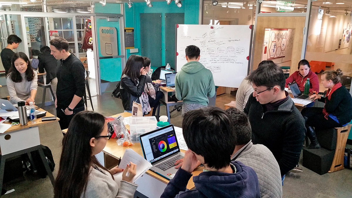 University students cluster in teams around desks with laptop computers. A dry erase board with a hand-drawn idea map stands in the background.