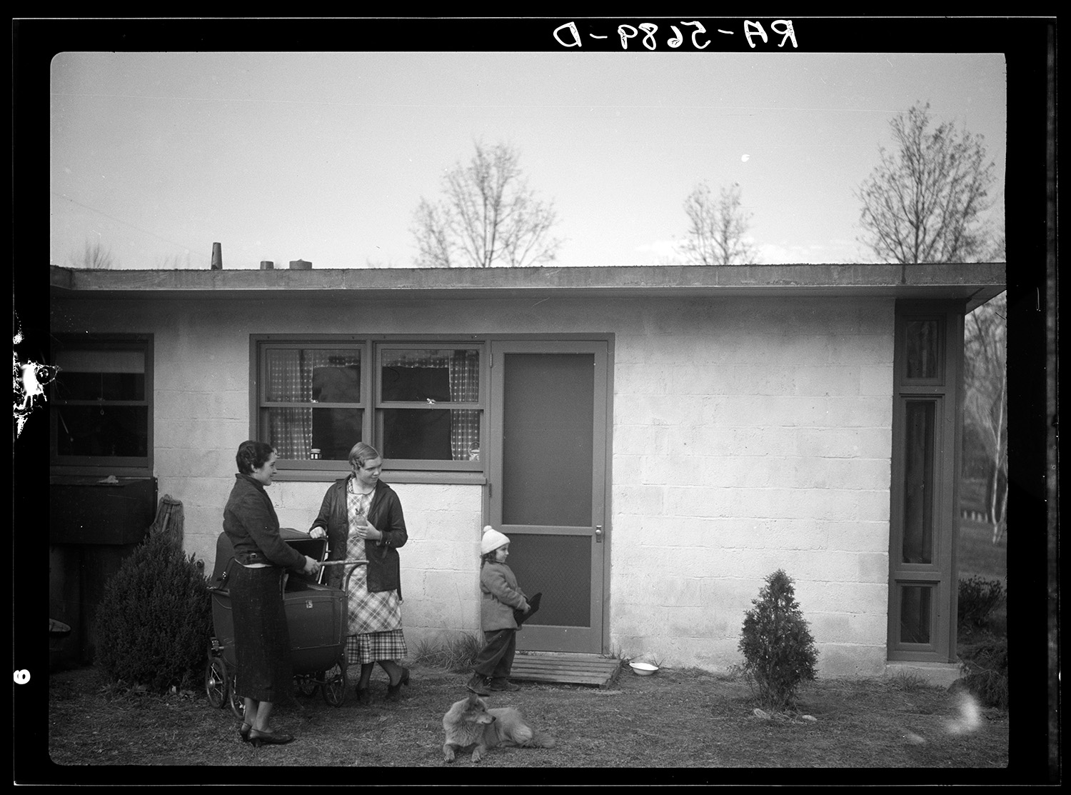 A black-and-white photo showing two women standing outside a modest house, with a young child and dog.