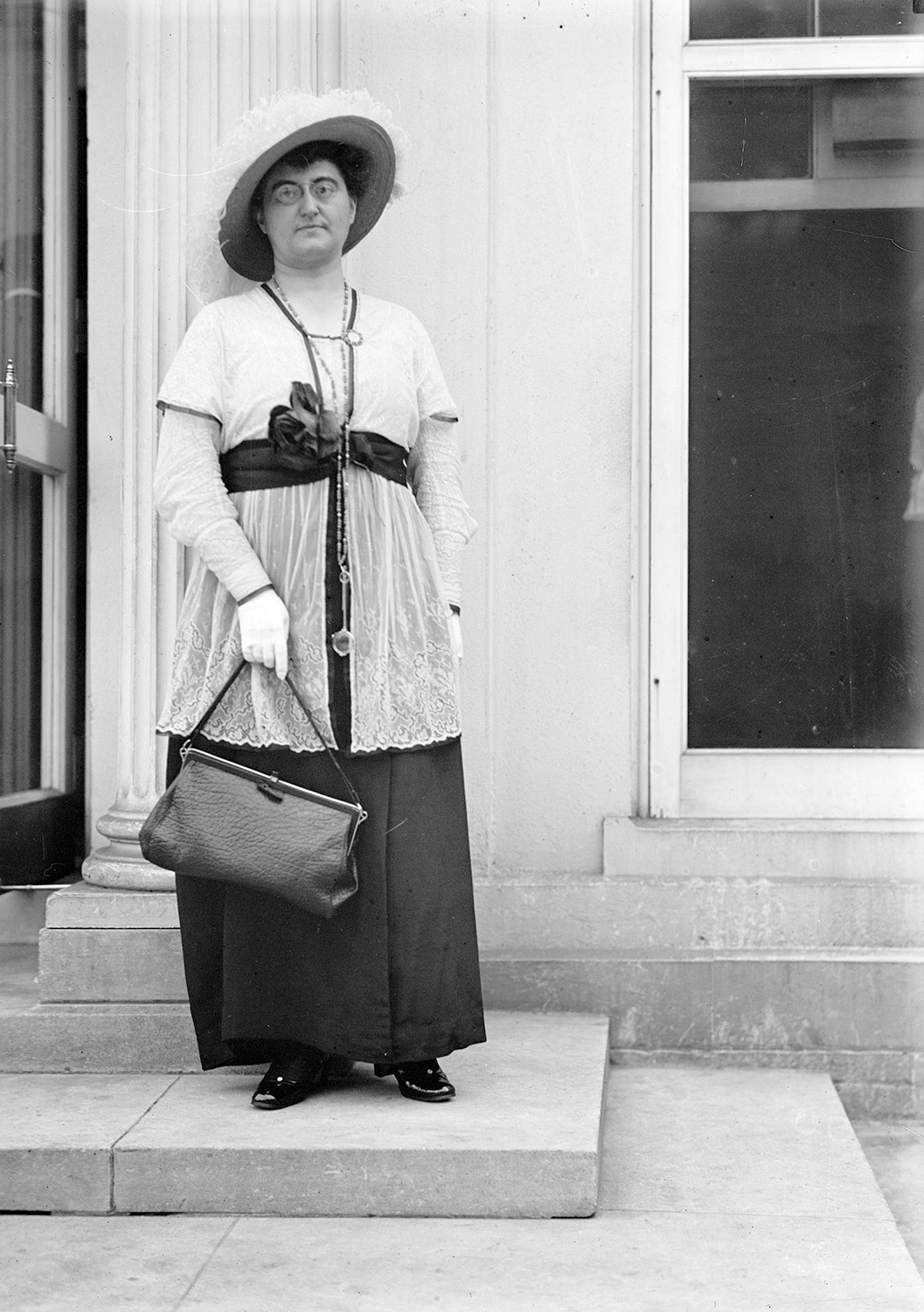 A portrait of Rosika Schwimmer, standing outside a building and facing the viewer. She is wearing a hat, a long black skirt with a white blouse, and white gloves.  She is holding a purse in her right hand.