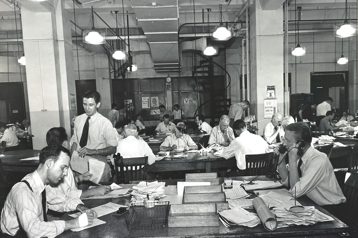 In a black and white photo of a newsroom, several editors and assistants work at desks.