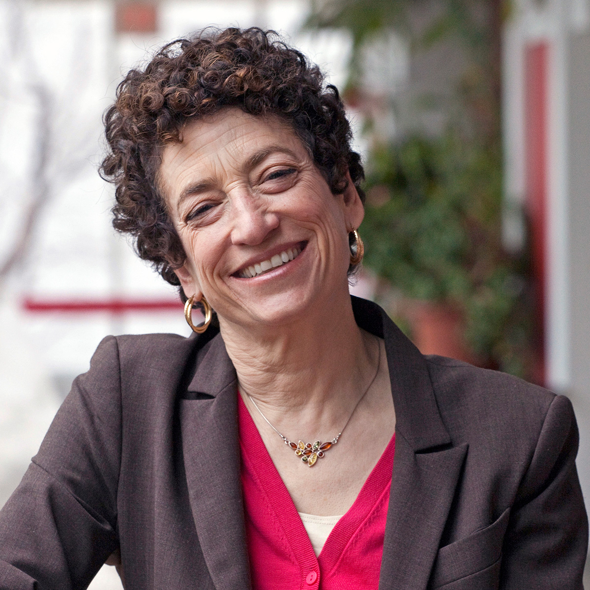 A photo of Naomi Oreskes, a light-skinned woman with dark curly hair. She is wearing a red sweater underneath a dark colored blazer and is smiling at the viewer.