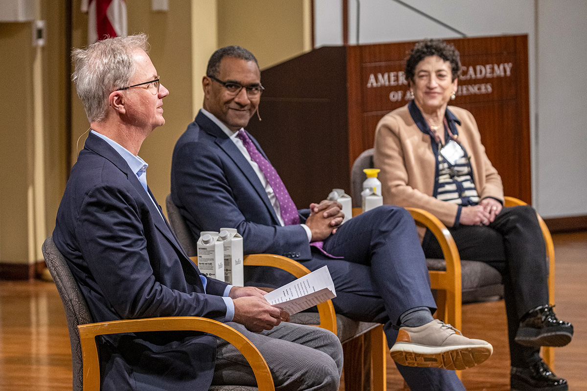 Three individuals are seated in chairs next to each other. Holden Thorp, on the left, has light skin, short gray hair, is wearing glasses, and is holding some papers in his hands. Sean Decatur, in the middle, has dark skin, is wearing a suit with a purple tie, and smiling toward the camera. Naomi Oreskes, on the right, has light skin, short curly hair, is wearing a beige blazer, and is smiling.