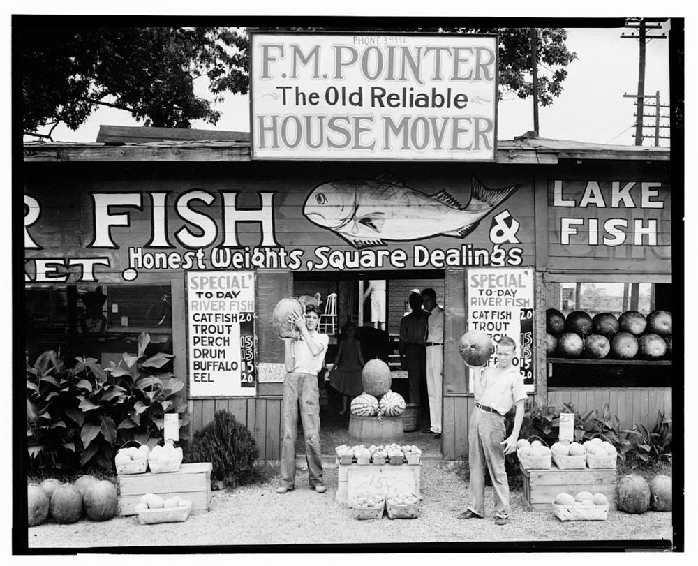Two youths standing in front of a store holding pumpkins. Other fruits and vegetables are on display. 