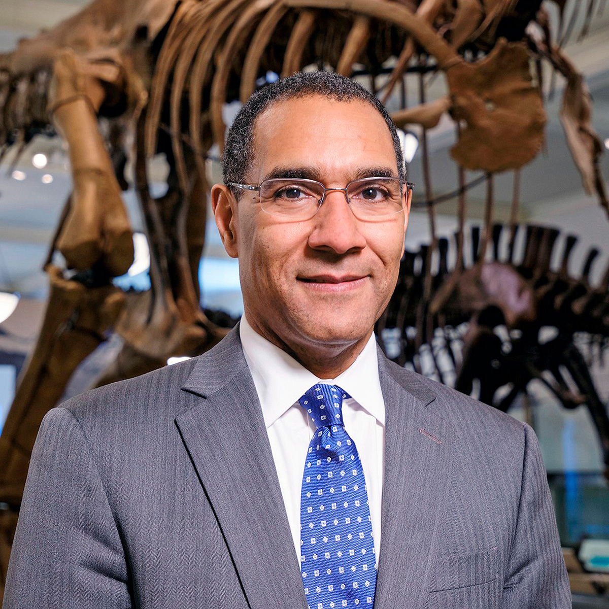 A photo of Sean Decatur, a dark-skinned man wearing glasses, a white shirt, blue tie, and a gray suit jacket. He is standing in front of a dinosaur skeleton in a museum exhibition space.  He is smiling at the viewer.