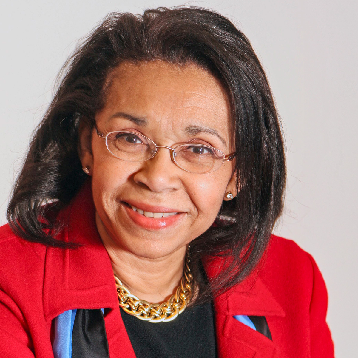 A photo of Shirley Malcom, a dark-skinned woman wearing glasses, a red jacket, and a black top accessorized with a gold chain necklace and a blue-and-black scarf. She is smiling at the camera.