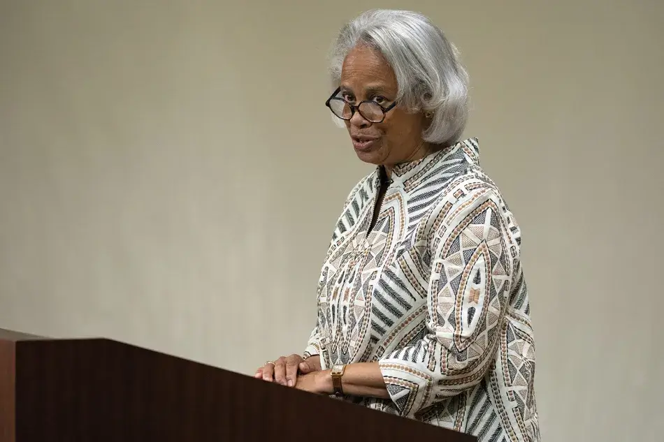 A photo of Ann Fudge, a woman with brown skin and white shoulder-length hair. She wears a white, black, and gold coat with the zipper on the collar. Photo by Martha Stewart Photography.