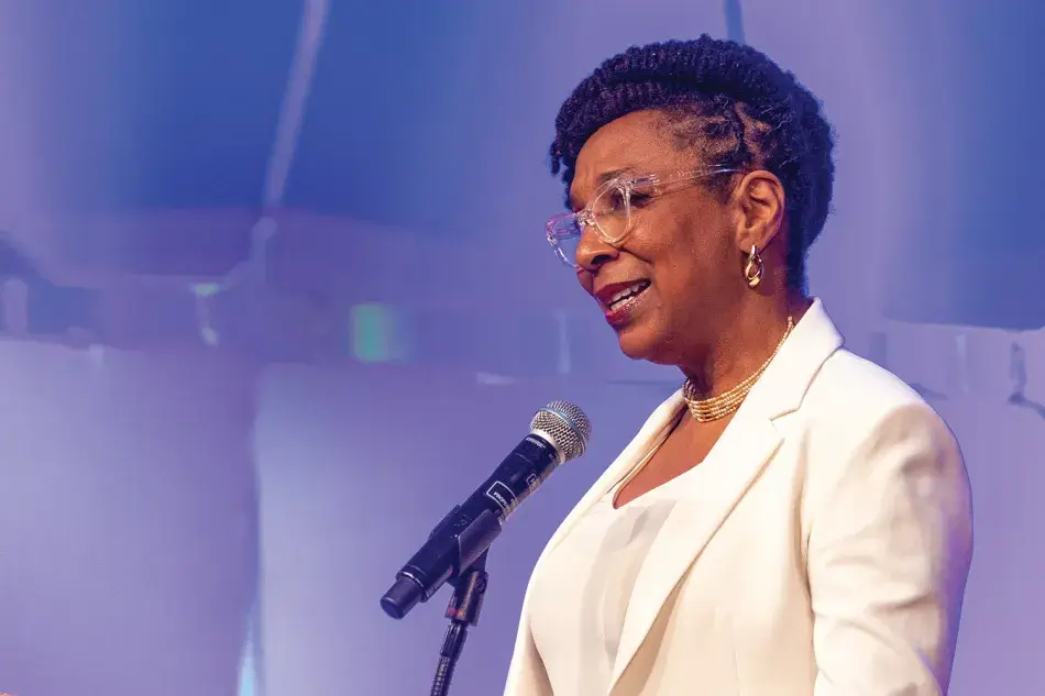 A photo of Kimberlé W. Crenshaw standing at a microphone at the 2022 Induction ceremony. She has brown skin and long black hair, worn in braids done up in an elegant twist. She wears a white suit, gold earrings, and a gold necklace. She smiles and addresses the audience. Photo by Michael DeStefano Photography.