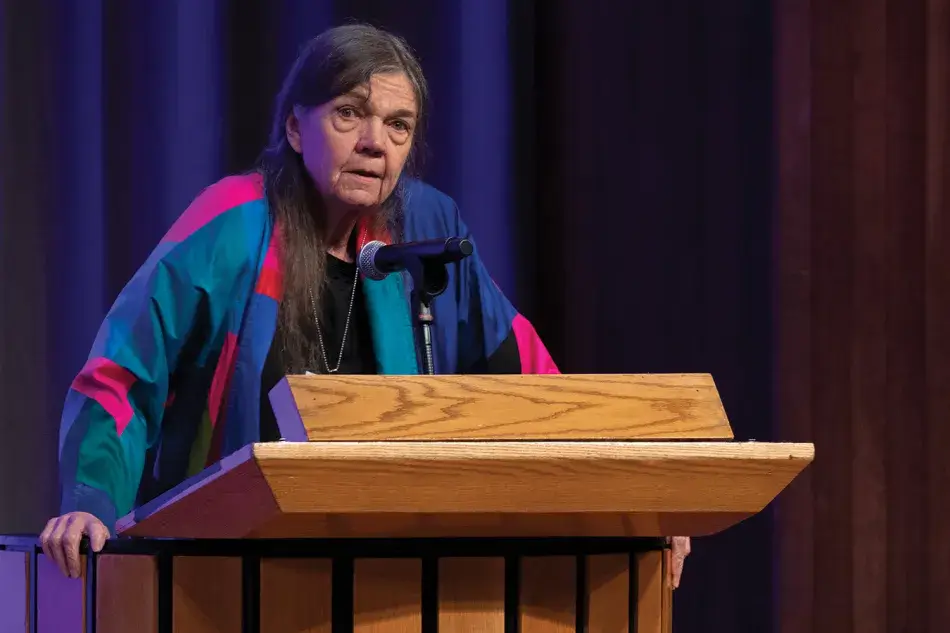 A photo of Patricia Limerick standing at a podium at the 2022 Induction ceremony. She has pale skin and long dark hair. She wears a multicolored suit, black blouse, and a silver necklace. She addresses the audience. Photo by Martha Stewart Photography.