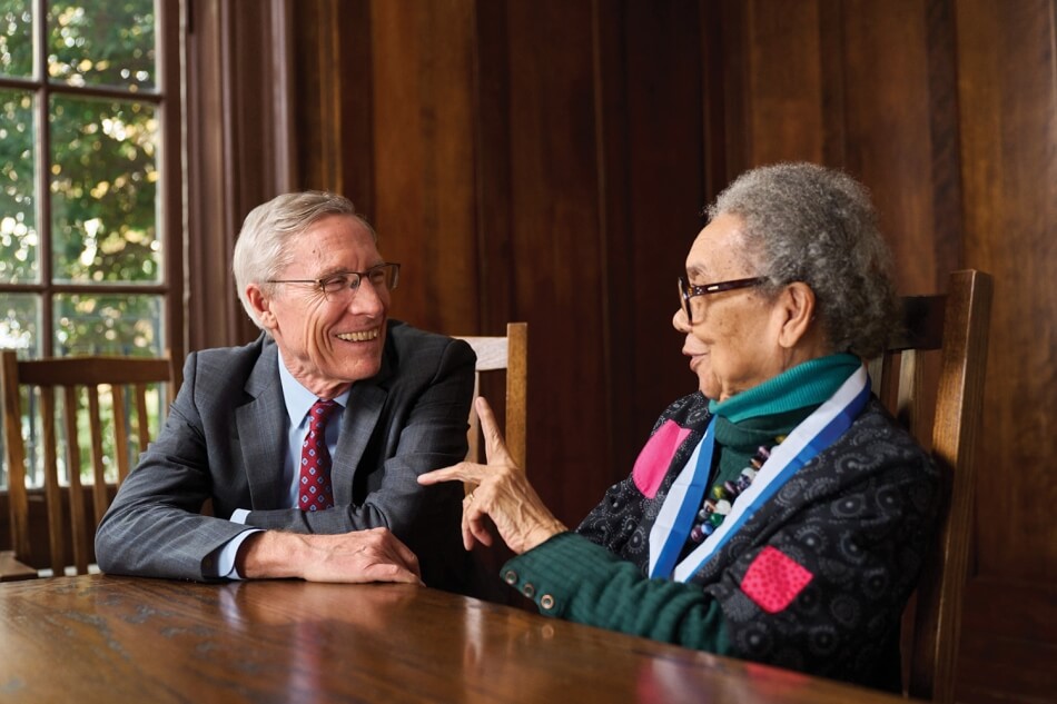 A photo of David Oxtoby and Marian Wright Edelman sitting side by side at a wooden table. Both smile, and Edelman holds one hand up as if in midspeech. Photo by Noah Willman.