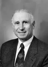 A black and white headshot of Arthur Gelb. He has short hair and wears a tweed suit with a white collared shirt and tie. He smiles gently at the viewer.