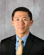 A headshot of Goodwin Liu, a person with brown skin, brown eyes, and short dark hair. He wears a suit, faces the viewer, and smiles.