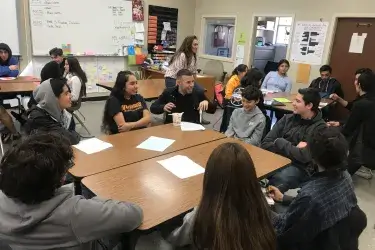 Candid photograph of a teacher sitting around a table and laughing with several high school English students