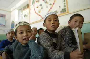 Six young students sit in a classroom in Gansu province, China. They look toward the front of the class. One student holds a book. The students have light brown skin and black hair. Many wear short white hats traditional among Salar people.