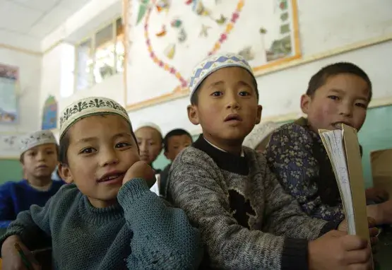 Six young students sit in a classroom in Gansu province, China. They look toward the front of the class. One student holds a book. The students have light brown skin and black hair. Many wear short white hats traditional among Salar people.