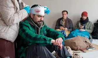 An adult sits on the floor beside a backpack. They have pale skin, a thick dark mustache, and short black hair. A person stands behind them and bandages the top of their head. Red can be seen through the bandages. Two other people wait in the background.