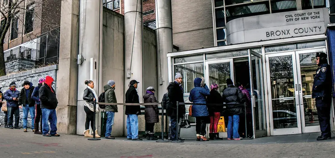 A line forms outside of the Bronx county housing court. People stand outside the court in jackets as a security guard watches the courthouse entrance.