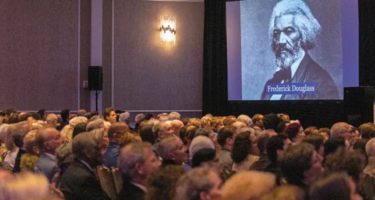 An audience seated in chairs looks at a projection of a black and white image of Frederick Douglass as an older man with white hair and a beard and wearing a white shirt and dark jacket and tie.