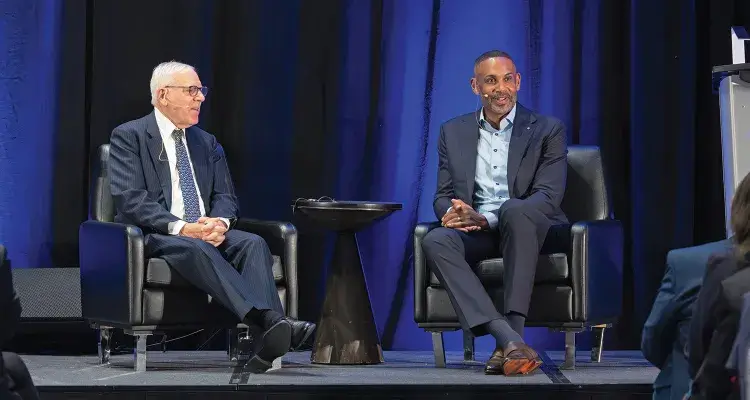 A photo of two people seated on a stage with a small round table between them.  On the left is David M. Rubenstein, a light-skinned man, wearing a dark suit and tie, who is looking at the other seated individual. On the right is Grant Hill, a dark-skinned man, wearing a business suit and looking out at the audience.